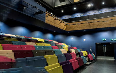 Hire Us, image is of our rainbow coloured double seating. The walls are a deep blue with lights beaming down.