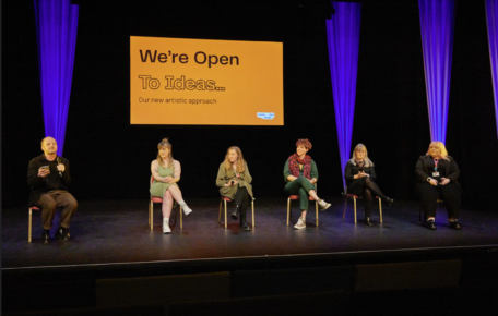 6 people sat on stage. Behind them is a orange slide which says the words: We're Open To Ideas.