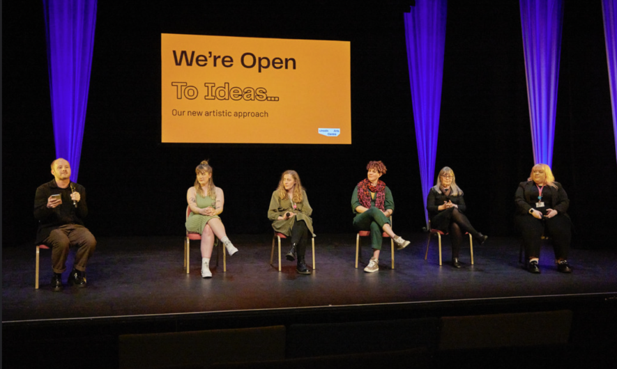6 people sat on stage. Behind them is a orange slide which says the words: We're Open To Ideas.