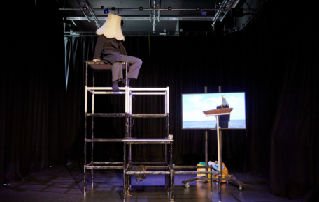 A performer sat on top of a scaffold with a lampshade on their head. There is a brush and a screen to the side of them.