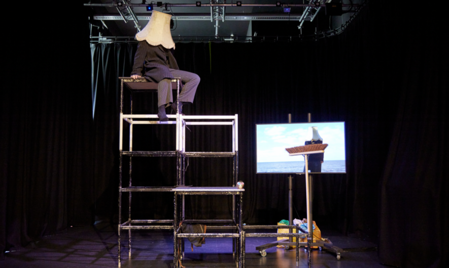 A performer sat on top of a scaffold with a lampshade on their head. There is a brush and a screen to the side of them.