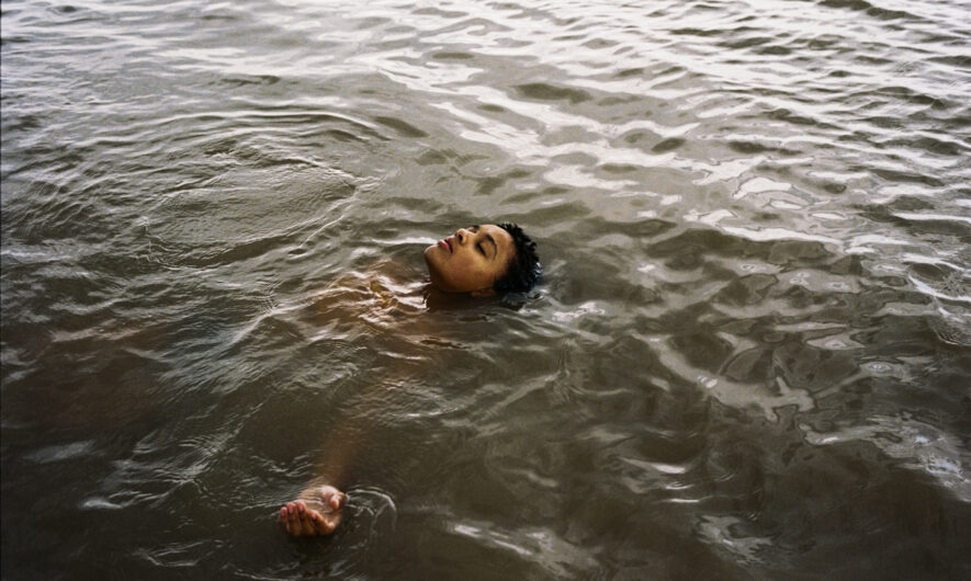 Young person floats in dark water with their face out of the water and eyes closed.