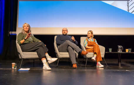 Three speakers are sat on stage in grey chairs and face the audience. They seem amidst a discussion or train of thought. The lady on the right wears a green jumper and has grey long hair. The man in the middle wears a blue jumper and glasses. The lady on the left wears a orange jumpsuit and has a blonde bob haircut.