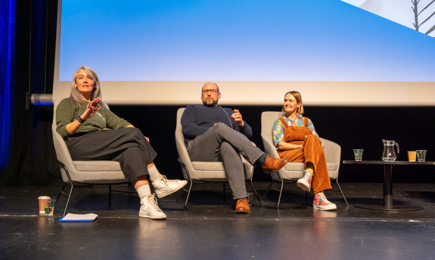 Three speakers are sat on stage in grey chairs and face the audience. They seem amidst a discussion or train of thought. The lady on the right wears a green jumper and has grey long hair. The man in the middle wears a blue jumper and glasses. The lady on the left wears a orange jumpsuit and has a blonde bob haircut.