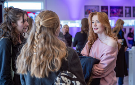 Three young women stand together. Two have their backs faced to the camera and one faces the camera. In the back there are other people blurred from focus that seem to be having conversations.