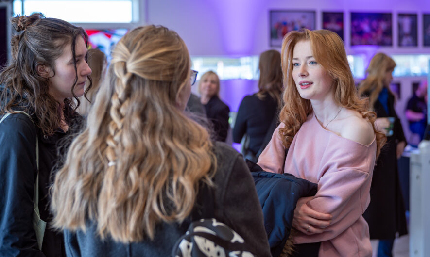 Three young women stand together. Two have their backs faced to the camera and one faces the camera. In the back there are other people blurred from focus that seem to be having conversations.