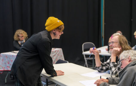 Woman lean over table to discuss with two seated participants during a creative workshop.