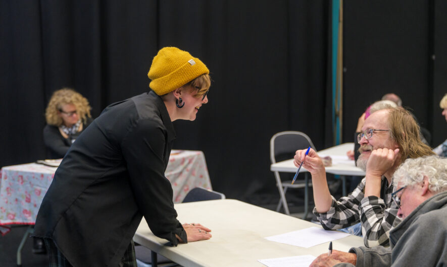 Woman lean over table to discuss with two seated participants during a creative workshop.