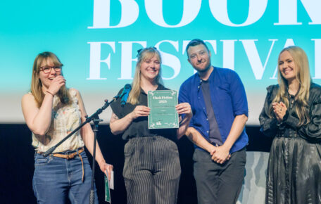Four people stand on stage smiling towards the camera, one is holding a Flash Fiction certificate.