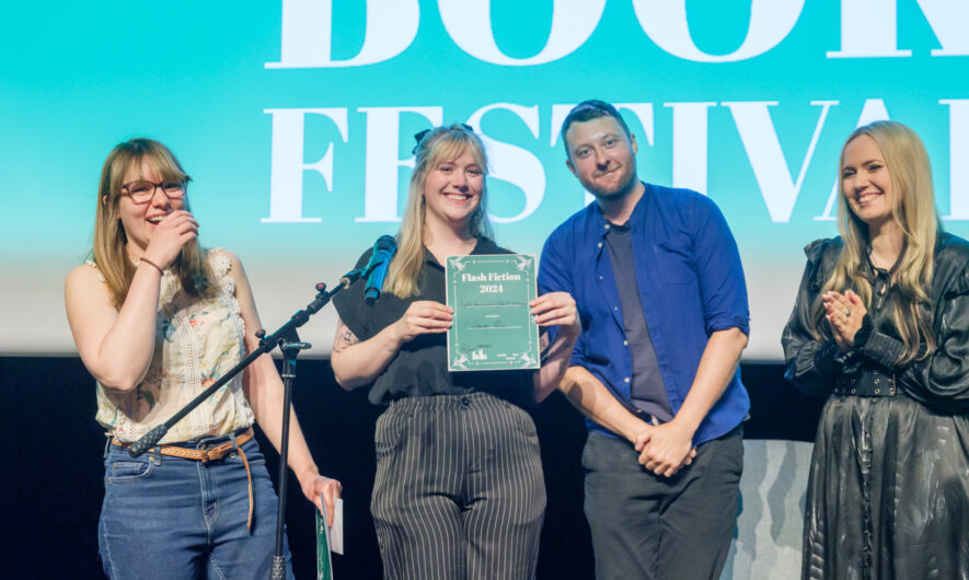 Four people stand on stage smiling towards the camera, one is holding a Flash Fiction certificate.