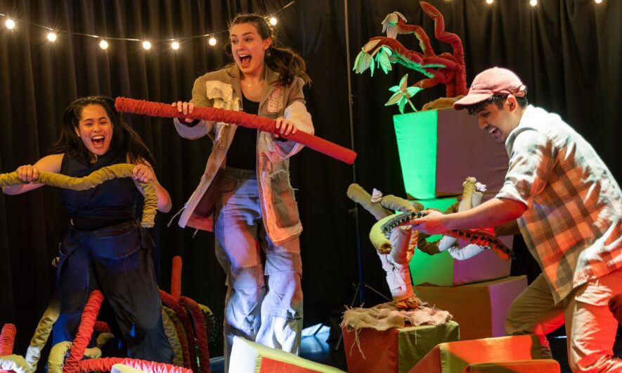 three performers on stage from 'Lose the path, find your breath'. They have excited expressions and are holding a large worm like object. The set has small fake plants and soft cubes stacked around.