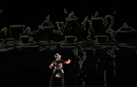 A man stands on stage with light shining from behind. He wears a tophat and tailcoat. The backdrop is very dark and overtop there is an outline of serval teapots and cups.