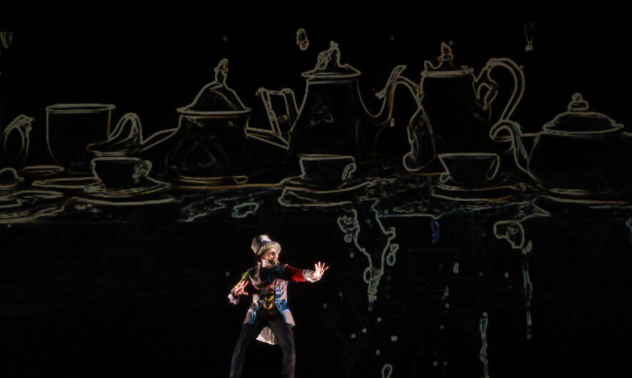 A man stands on stage with light shining from behind. He wears a tophat and tailcoat. The backdrop is very dark and overtop there is an outline of serval teapots and cups.