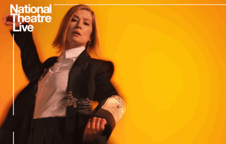 Rosamund Pike poses against a orange background with her right arm stretched up into the air. She wears a black suit with a white shirt and has a blond bob hair style. Inter Alia is written in capitol letters on the background.