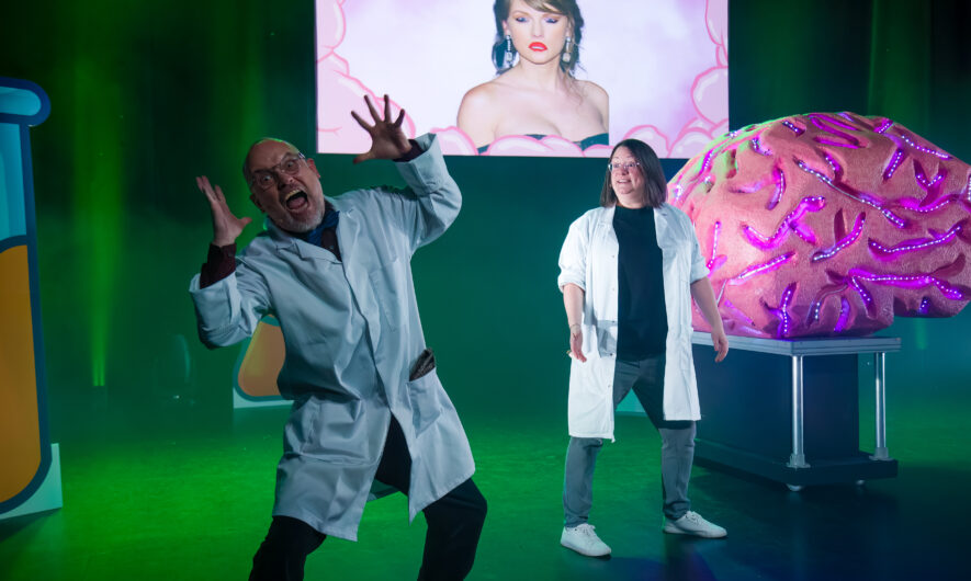 A man on a woman stand on stage. The man infront has both his arms raised into the air and has a screaming expression on his face. The lady in the back is smiling. They both wear white lab coats with black t-shirts and trousers. There is a illuminated prop brain covered with lights and a projector shows Tayler Swift with her face upside down.