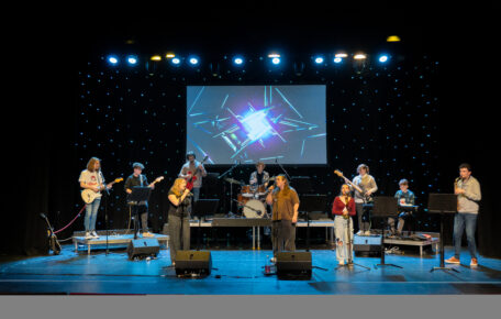 ten students are stood on stage with different instruments. Guitars, a clarinet, drum set and three singers at the front. The background is lit up with small LED light as if they were stars.