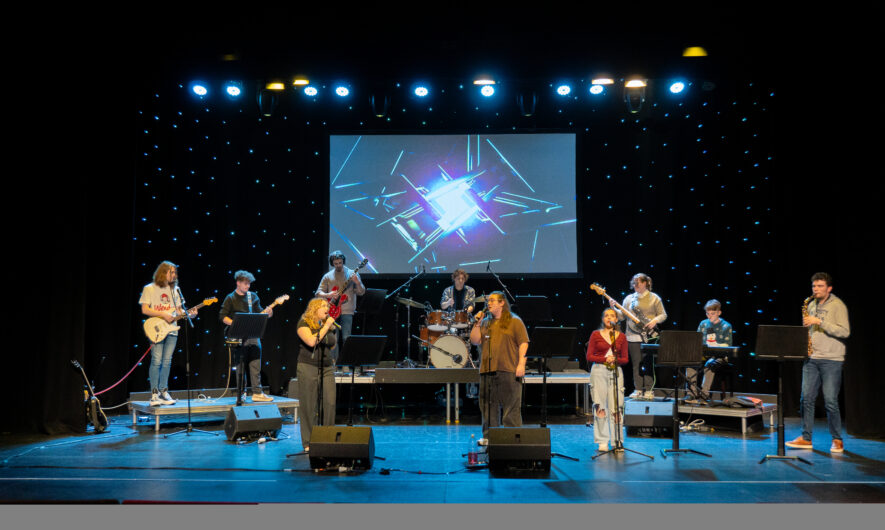 ten students are stood on stage with different instruments. Guitars, a clarinet, drum set and three singers at the front. The background is lit up with small LED light as if they were stars.