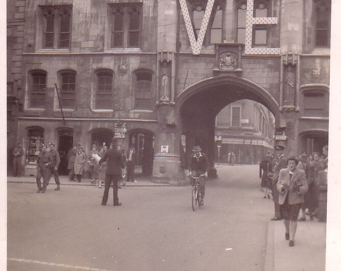 Old black and white photograph depicting the Guildhall curing VE celebrations. The Guildhall building has a big VE sign hung across its central windows with people walking through its arches.